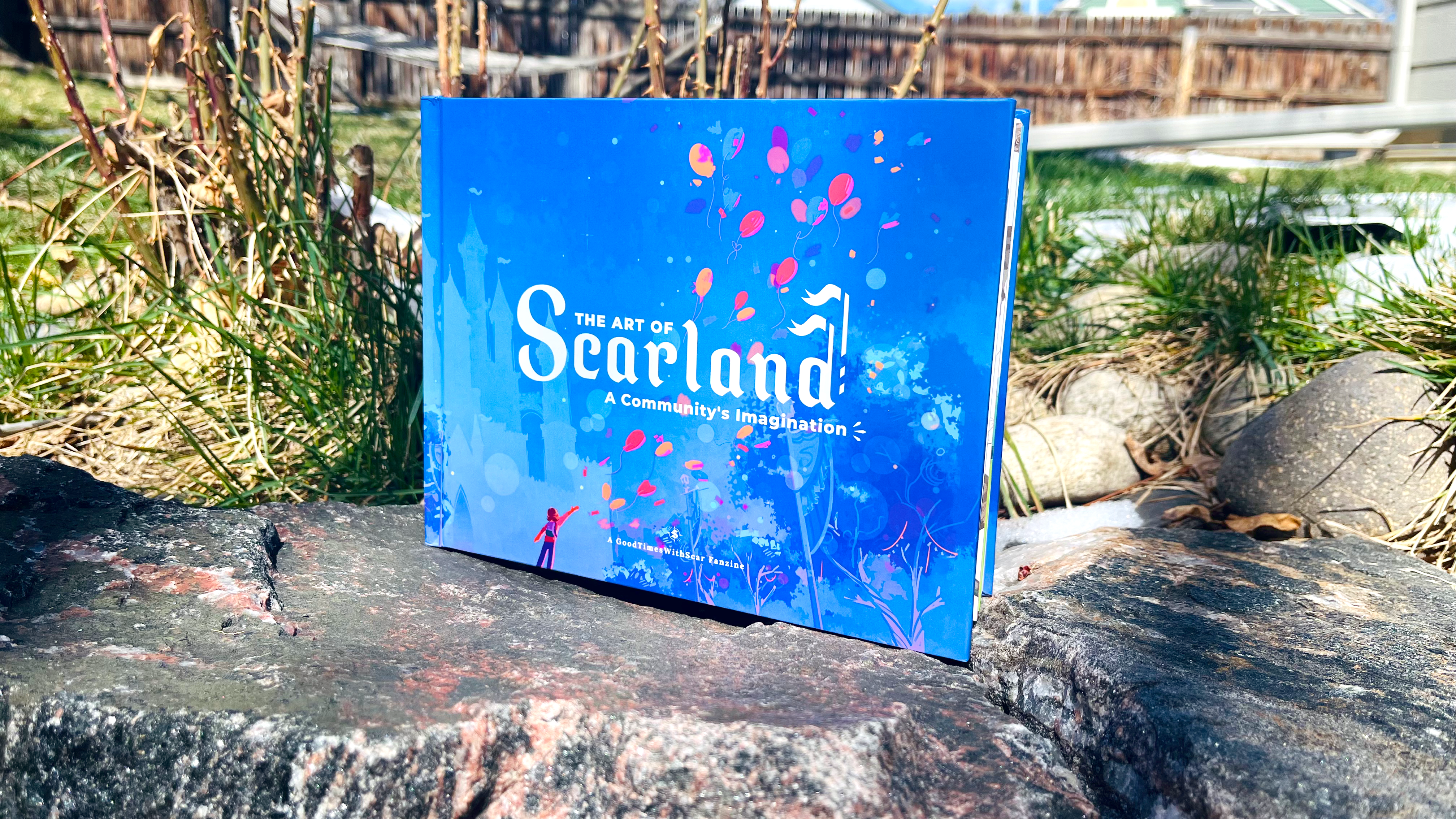 Photo of the Scarland Artbook outdoors. Behind it is grass and a rosebush, and it sits on blue rocks. A fence is distantly visible in the background.